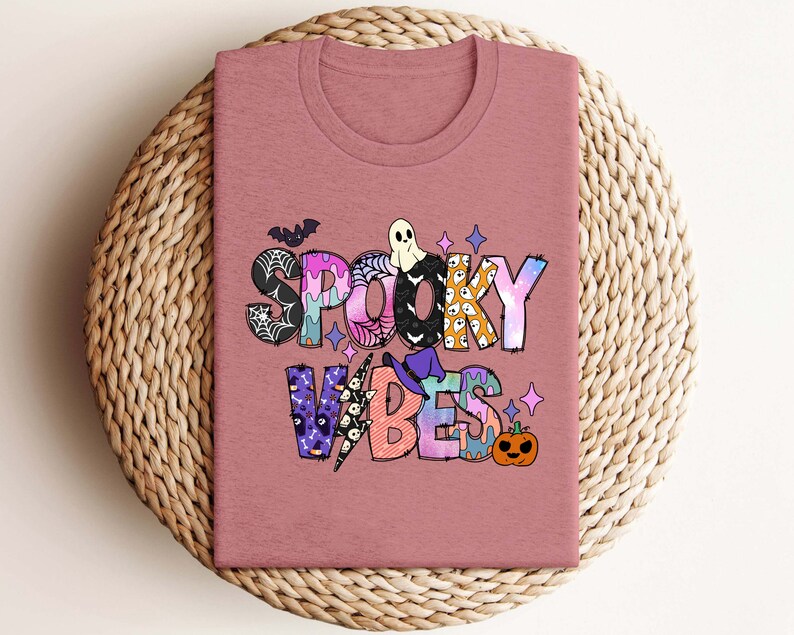 May include: A pink t-shirt with a colorful graphic that says "Spooky Vibes". The graphic includes a ghost, a witch's hat, a spiderweb, and a pumpkin.