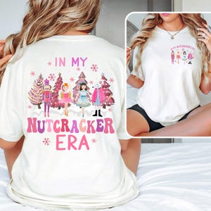 May include: Cream-colored t-shirt with a graphic featuring a Nutcracker theme. The design includes pink and purple text that reads "IN MY NUTCRACKER ERA" and illustrations of Nutcracker characters and Christmas trees. A smaller version of the design is on the front.