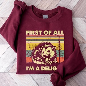 Może przedstawiać: Bordowa bluza z retro grafiką oposa z tekstem "First of all I'm a Delig".