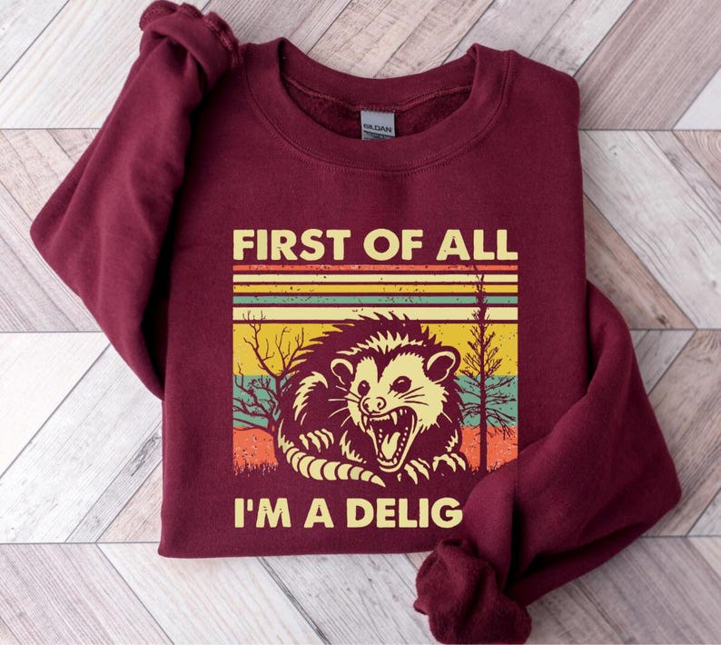 Może przedstawiać: Bordowa bluza z retro grafiką oposa z tekstem "First of all I'm a Delig".