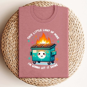 May include: A pink t-shirt featuring a graphic of a green wheelie bin on fire with a smiling face and the text "This little light of mine I'm gonna let it shine".