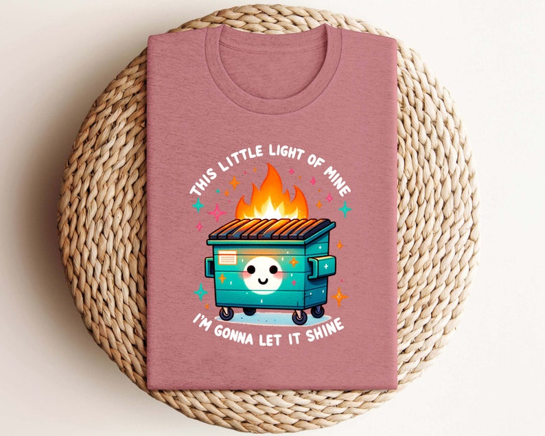 May include: A pink t-shirt featuring a graphic of a green wheelie bin on fire with a smiling face and the text "This little light of mine I'm gonna let it shine".