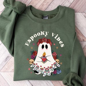 May include: Olive green sweatshirt with the text "Espooky Vibes" and a ghost graphic. The ghost wears a floral crown and holds a heart with a flame. The design includes roses, skulls, and stars, creating a festive Halloween theme.