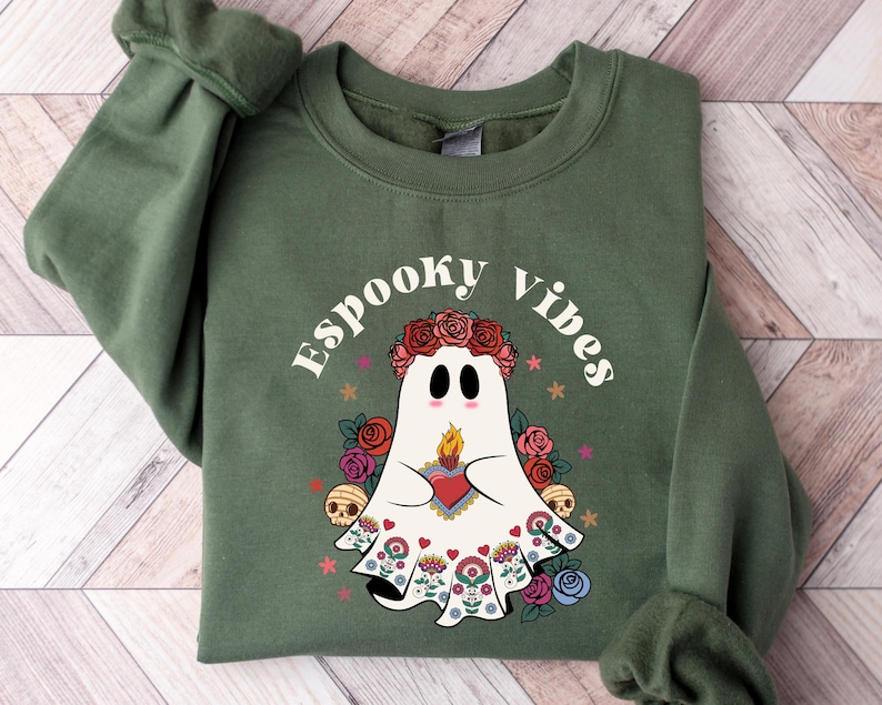 May include: Olive green sweatshirt with the text "Espooky Vibes" and a ghost graphic. The ghost wears a floral crown and holds a heart with a flame. The design includes roses, skulls, and stars, creating a festive Halloween theme.