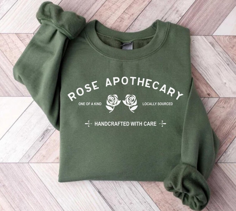 Puede incluir: Una sudadera verde con el texto "ROSE APOTHECARY" en blanco. El texto est&aacute; rodeado por dos rosas blancas y las palabras "ONE OF A KIND" y "LOCALLY SOURCED". Debajo del texto est&aacute; la frase "HANDCRAFTED WITH CARE".