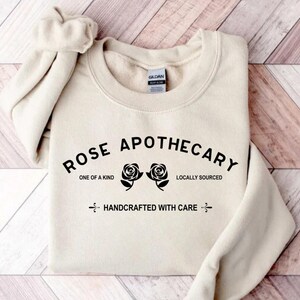 Puede incluir: Una sudadera de color crema con el texto "ROSE APOTHECARY" en letras negras. El texto est&aacute; arqueado sobre dos ilustraciones de rosas negras. Debajo de las rosas, el texto dice "ONE OF A KIND" y "LOCALLY SOURCED". Debajo de eso, el texto dice "HANDCRAFTED WITH CARE".