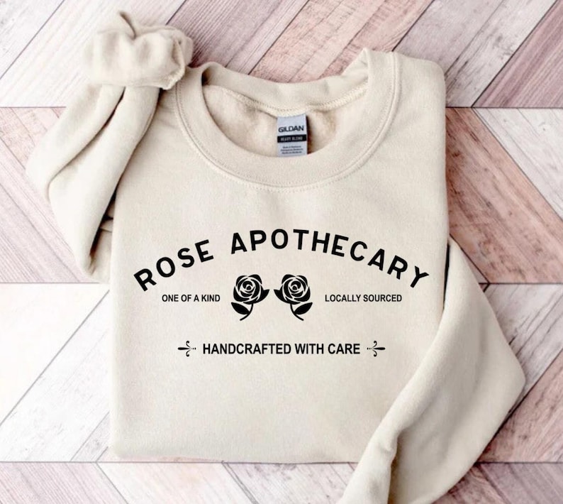 Puede incluir: Una sudadera de color crema con el texto "ROSE APOTHECARY" en letras negras. El texto est&aacute; arqueado sobre dos ilustraciones de rosas negras. Debajo de las rosas, el texto dice "ONE OF A KIND" y "LOCALLY SOURCED". Debajo de eso, el texto dice "HANDCRAFTED WITH CARE".
