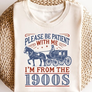 Pu&ograve; includere: Maglietta bianca con un design grafico blu e rosso. Il grafico presenta una carrozza trainata da un cavallo con il testo "Per favore, sii paziente con me, sono degli anni 1900".