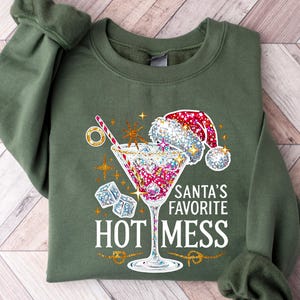 May include: Olive green sweatshirt with a festive graphic. The design features a cocktail glass with a Santa hat, ice cubes, and the text "Santa's Favorite HOT MESS" in white. Gold accents and starbursts complete the holiday theme.