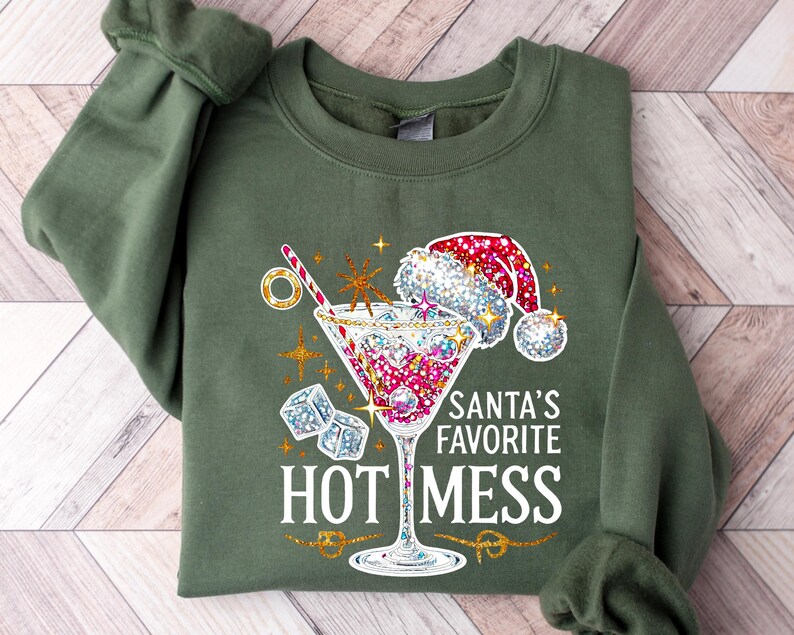 May include: Olive green sweatshirt with a festive graphic. The design features a cocktail glass with a Santa hat, ice cubes, and the text "Santa's Favorite HOT MESS" in white. Gold accents and starbursts complete the holiday theme.