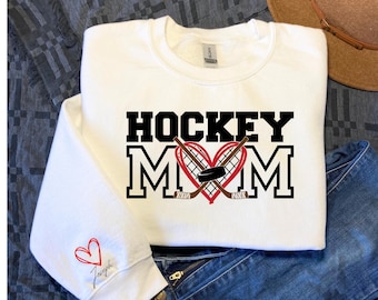 May include: White sweatshirt with the text "HOCKEY MOM" in black letters. A red heart with black hockey sticks and a puck is in the middle of the text.