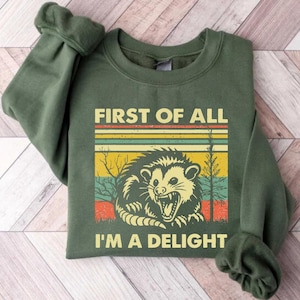 Może przedstawiać: Zielona bluza z nadrukiem w stylu retro przedstawiającym oposa z tekstem "First of all I'm a delight".