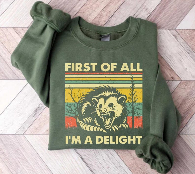 Może przedstawiać: Zielona bluza z nadrukiem w stylu retro przedstawiającym oposa z tekstem "First of all I'm a delight".