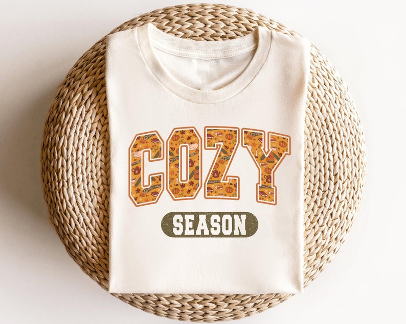 May include: A white t-shirt with a graphic print of the word "COZY" in orange and brown with a fall-themed pattern. The text "SEASON" is printed in green below the word "COZY".