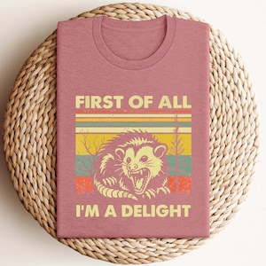 Może przedstawiać: R&oacute;żowy t-shirt z grafiką w stylu vintage przedstawiającą oposa z tekstem "First of all I'm a delight".