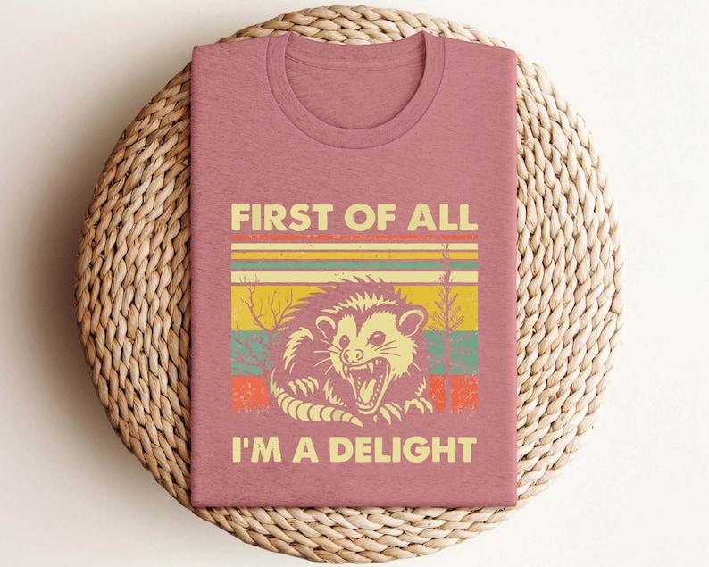 Może przedstawiać: R&oacute;żowy t-shirt z grafiką w stylu vintage przedstawiającą oposa z tekstem "First of all I'm a delight".
