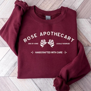 Puede incluir: Sudadera color burdeos con texto blanco que dice "Rose Apothecary" con dos rosas blancas y el texto "One of a Kind", "Locally Sourced" y "Handcrafted with Care".