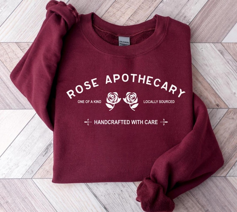 Puede incluir: Sudadera color burdeos con texto blanco que dice "Rose Apothecary" con dos rosas blancas y el texto "One of a Kind", "Locally Sourced" y "Handcrafted with Care".