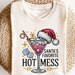 May include: Cream-colored t-shirt featuring a festive cocktail design. The graphic includes a martini glass with a Santa hat, ice cubes, and the text "SANTA'S FAVORITE HOT MESS" in black and gold. The design is embellished with glitter.