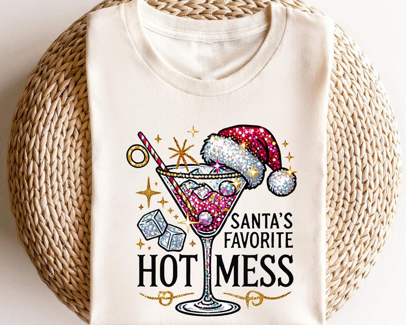May include: Cream-colored t-shirt featuring a festive cocktail design. The graphic includes a martini glass with a Santa hat, ice cubes, and the text "SANTA'S FAVORITE HOT MESS" in black and gold. The design is embellished with glitter.