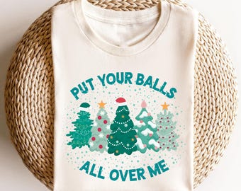 以下が含まれることがあります： クリスマスをテーマにしたデザインの、オフホワイトのTシャツ。緑、青、ピンクの色合いで装飾された5本のクリスマスツリーが描かれています。「PUT YOUR BALLS ALL OVER ME」というテキストがターコイズ色で表示されています。ツリーはオーナメントとサンタの帽子で飾られています。