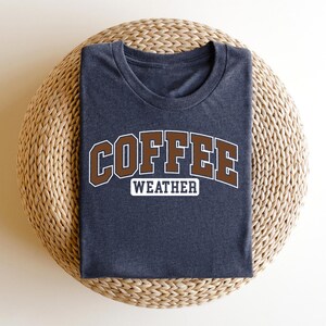 K&ouml;nnte beinhalten: Ein dunkelgraues T-Shirt mit dem Text "COFFEE WEATHER" in braunen Buchstaben mit wei&szlig;em Umriss.