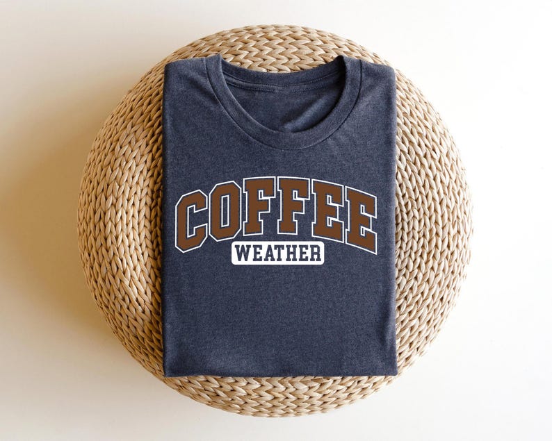 K&ouml;nnte beinhalten: Ein dunkelgraues T-Shirt mit dem Text "COFFEE WEATHER" in braunen Buchstaben mit wei&szlig;em Umriss.