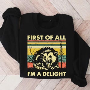 Może przedstawiać: Czarny T-shirt z długim rękawem z retro grafiką oposa w leśnym otoczeniu. Na koszulce nadrukowany jest tekst "First of all, I'm a delight".
