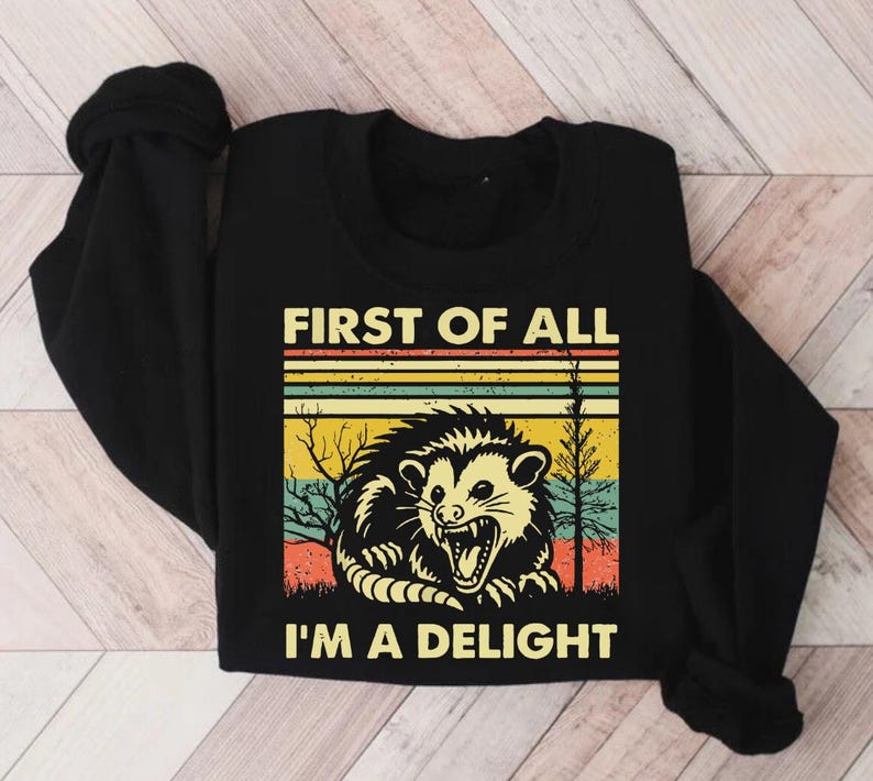 Może przedstawiać: Czarny T-shirt z długim rękawem z retro grafiką oposa w leśnym otoczeniu. Na koszulce nadrukowany jest tekst "First of all, I'm a delight".