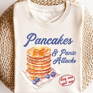 May include: A white t-shirt with a vintage-style graphic of a stack of pancakes with butter and syrup, blueberries, and the text "Pancakes & Panic Attacks" in blue. The text "buy one get one" is in red.