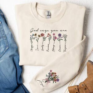 May include: A beige sweatshirt with the text "God says you are" and a floral design. The words "Unique, Special, Lovely, Precious, Strong, Chosen, Forgiven" are listed below the flowers.
