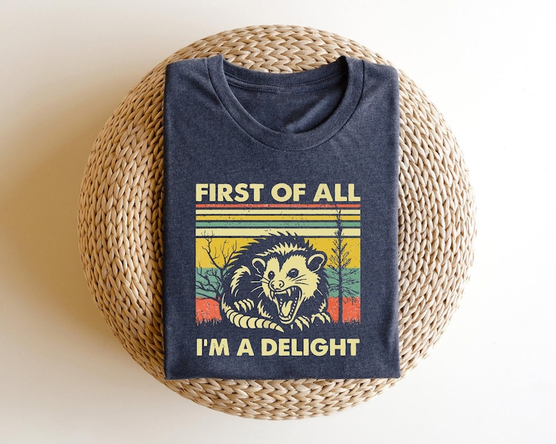 Może przedstawiać: Koszulka w ciemnoniebieskim kolorze z grafiką vintage przedstawiającą oposa. Grafika zawiera tekst "First of all, I'm a delight" w kolorystyce tęczy.