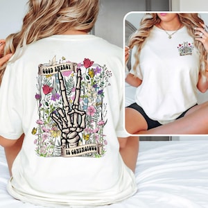May include: White t-shirt with a floral design and a skeleton hand making a peace sign. The text "Good Energy is Contagious" is printed on the shirt.