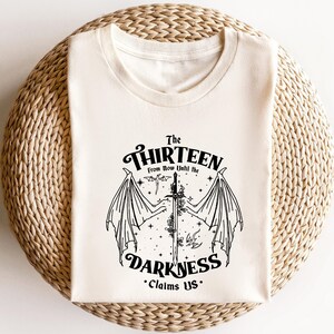 May include: A white t-shirt with a black graphic design featuring a sword with wings and the text "The Thirteen From Now Until the Darkness Claims Us."