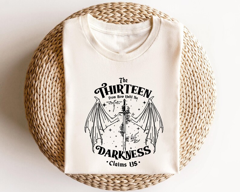 May include: A white t-shirt with a black graphic design featuring a sword with wings and the text "The Thirteen From Now Until the Darkness Claims Us."