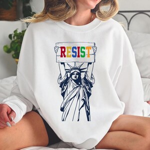 May include: White sweatshirt with a graphic of the Statue of Liberty holding a sign that says "RESIST" in rainbow colors.