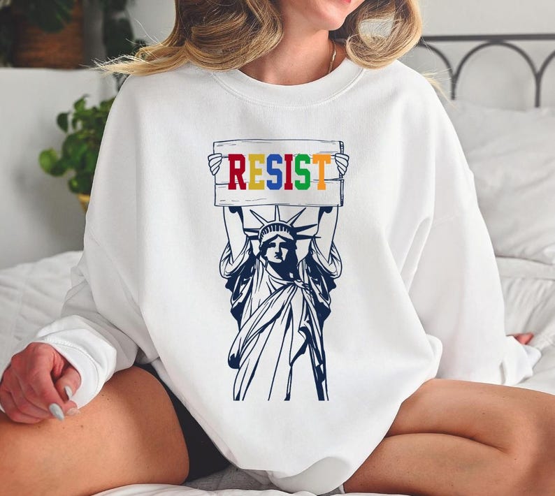 May include: White sweatshirt with a graphic of the Statue of Liberty holding a sign that says "RESIST" in rainbow colors.
