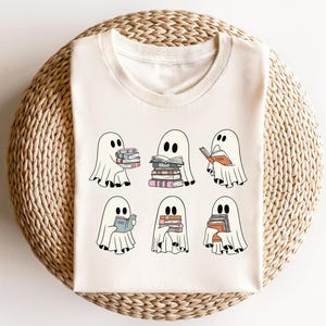 May include: A white t-shirt with a design of six ghosts holding or reading books. The ghosts are all different sizes and have different expressions. The text "Bookish Ghosts" is printed on the shirt.