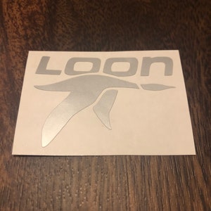May include: A silver sticker featuring the word "LOON" and a bird graphic. The sticker is on a white background, set against a dark wood grain surface. The font is a sans-serif style.