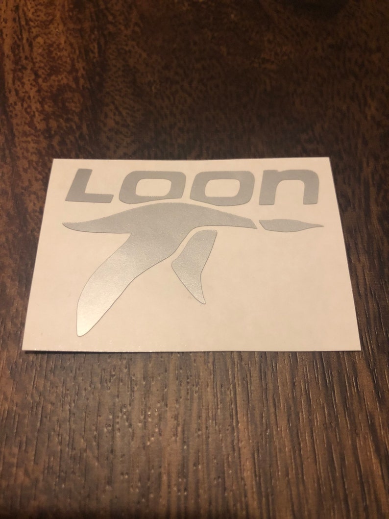 May include: A silver sticker featuring the word "LOON" and a bird graphic. The sticker is on a white background, set against a dark wood grain surface. The font is a sans-serif style.
