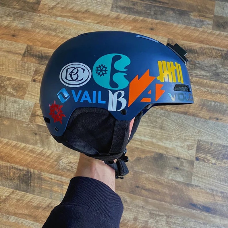 May include: A navy blue ski helmet with various colourful stickers, including the word "VAIL" and other graphic designs. The helmet has a black chin strap and a small camera mount on top. Designed for winter sports.