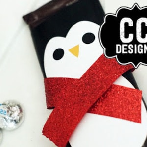 May include: A chocolate bar wrapped in a white penguin design with a red glitter scarf. The penguin has black eyes and a yellow beak. The chocolate bar is wrapped in black paper with the text "CC DESIGNS" in white.
