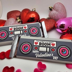 Op de afbeelding: Twee chocolade repen verpakt in zilverfolie met een Valentijnsdagthema. De wikkels hebben een boombox-ontwerp met de tekst "You make my heart BOOM" en "Happy Valentine's Day".