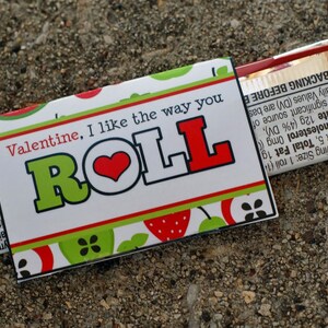 May include: A Valentine's Day card with a red heart and the text "Valentine, I like the way you ROLL" on a white background with green and red apple designs. The card is attached to a chocolate bar.