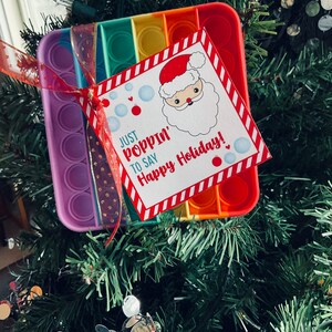May include: A rainbow pop-it toy with a red and white striped tag attached. The tag has a cartoon image of Santa Claus and the text "Just poppin' to say Happy Holidays!"