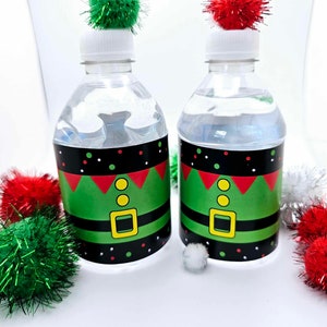 May include: Two clear plastic water bottles with green, red, black, and yellow elf costume labels. The bottles are topped with red and green fuzzy pom-poms.