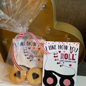 Op de afbeelding: Een Valentijnsdag-cadeauzak met een rood-wit gestreept lint. De tas bevat twee koekjes met hartvormige centra en twee kaarten met de tekst "Ik vind het leuk hoe je rolt" en een rolstoelsticker.