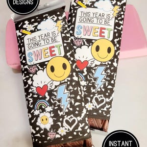 Può includere: Due barrette di cioccolato avvolte in carta a motivi bianchi e neri con la scritta "THIS YEAR IS GOING TO BE SWEET". Le confezioni presentano faccine sorridenti, cuori, arcobaleni e fulmini. L'immagine include anche il testo "CC DESIGNS" e "INSTANT DOWNLOAD".