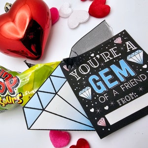 May include: A black card with the text "You're a GEM of a friend" with a diamond design and glitter. The card is tied with a black ribbon to a blue diamond-shaped paper cutout and a green and white candy wrapper.