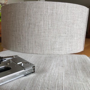 May include: A silver staple gun with a black handle, lying on a light grey fabric surface. The fabric has a woven texture.